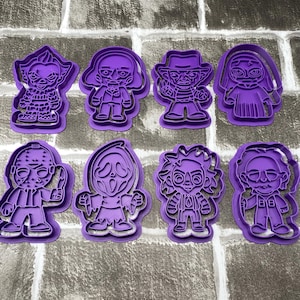 May include: A collection of eight purple plastic cookie cutters featuring iconic horror movie characters. The cutters include designs of a clown, a zombie, Freddy Krueger, a vampire, Jason Voorhees, Ghostface, and others.