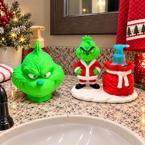 Peut inclure: Un distributeur de savon vert vif en forme de Grinch avec une pompe dorée, accompagné d'une figurine du Grinch en costume de Père Noël rouge et blanc à côté d'un distributeur en forme de sac rouge avec une pompe bleue sur socle blanc.