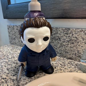 Peut inclure: Un distributeur de savon repr&eacute;sentant une figurine avec un masque blanc, des cheveux bruns et une combinaison bleue. La figurine tient un petit couteau argent&eacute; dans une main. Une pompe transparente est fix&eacute;e sur le flacon de savon violet.