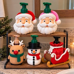 Peut inclure: Cinq distributeurs de savon sur le thème des fêtes sur une étagère en bois à deux niveaux. Deux têtes de Père Noël avec des bonnets rouge et rose, un renne beige avec une écharpe verte, un bonhomme de neige et un traîneau rouge.