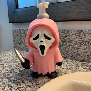 Peut inclure: Un distributeur de savon rose en forme de personnage encapuchonn&eacute; avec un masque blanc aux d&eacute;coupes noires pour les yeux et la bouche. Le personnage porte une robe rose, des gants noirs, des chaussures noires et tient un couteau argent&eacute;.