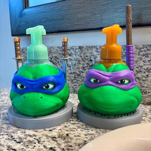 Peut inclure: Deux distributeurs de savon en forme de tortue verte sur des bases circulaires grises. Le distributeur de gauche a un masque bleu et deux épées miniatures, avec une pompe vert clair. Celui de droite a un masque violet et un bâton marron.