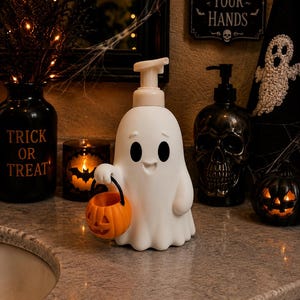 Peut inclure: Un distributeur de savon en forme de fant&ocirc;me blanc avec une pompe, arborant un visage souriant et tenant un petit seau citrouille orange en plastique avec une anse noire. Le distributeur est en mat&eacute;riau lisse et pos&eacute; sur une surface plane.