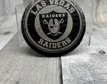 Puede incluir: Un posavasos circular de pizarra negra con el logotipo de los Las Vegas Raiders grabado en el centro. El diseño incluye el nombre del equipo arqueado sobre y debajo del emblema de escudo, que muestra a un jugador con casco y parche.