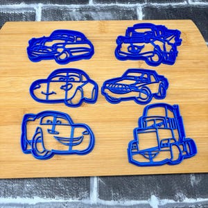 May include: Six blue plastic car-shaped cookie cutters on a wooden cutting board. The cutters include various car designs, such as a sedan, a race car, and a truck. The cutters are ideal for making car-themed cookies.