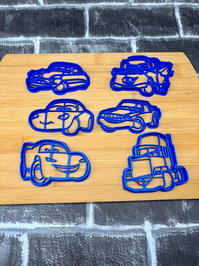May include: Six blue plastic car-shaped cookie cutters on a wooden cutting board. The cutters include various car designs, such as a sedan, a race car, and a truck. The cutters are ideal for making car-themed cookies.