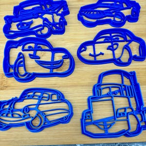 May include: Six blue plastic car-shaped cookie cutters are arranged on a wooden surface. The cutters vary in design, with some resembling cartoon cars and others more realistic vehicles. The cutters are made of a solid blue plastic.