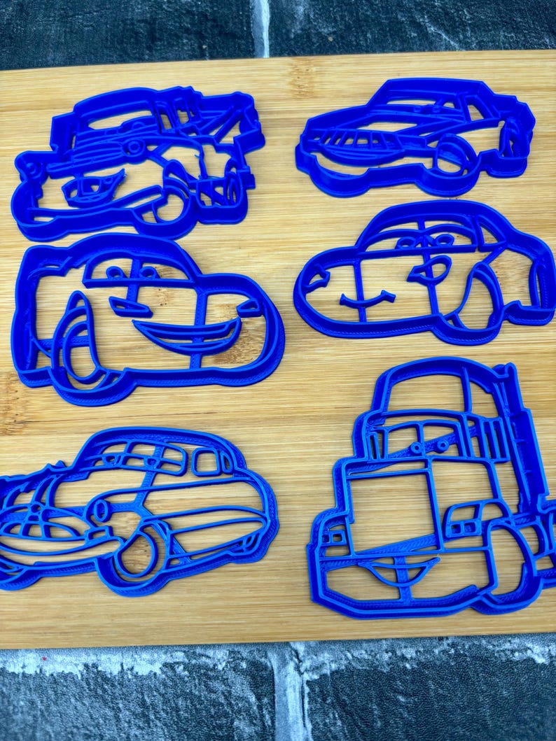 May include: Six blue plastic car-shaped cookie cutters are arranged on a wooden surface. The cutters vary in design, with some resembling cartoon cars and others more realistic vehicles. The cutters are made of a solid blue plastic.