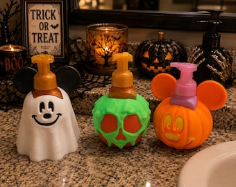 May include: Three decorative soap dispensers featuring Halloween themes. One is a white ghost with black ears, one is an orange apple with green slime, and one is an orange pumpkin with orange ears and a pink pump. Each has a functional soap pump.
