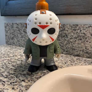 Peut inclure: Un distributeur de savon en forme de personnage portant un masque de hockey blanc avec des accents rouges. La figurine porte une veste verte, un pantalon gris, des chaussures noires et tient un petit couteau argent&eacute;. Une pompe orange est fix&eacute;e au sommet.
