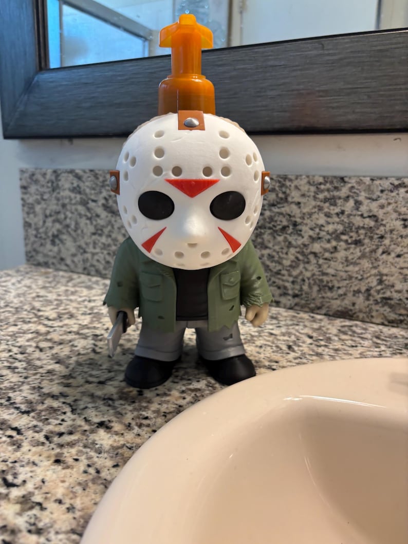 Peut inclure: Un distributeur de savon en forme de personnage portant un masque de hockey blanc avec des accents rouges. La figurine porte une veste verte, un pantalon gris, des chaussures noires et tient un petit couteau argent&eacute;. Une pompe orange est fix&eacute;e au sommet.