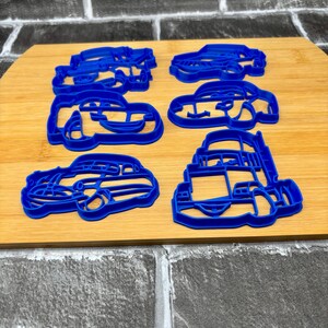 May include: Six blue plastic car-shaped cookie cutters on a wooden surface. The cutters are in various car designs, including a sedan, a convertible, and a truck. The cutters are made of a durable plastic material.