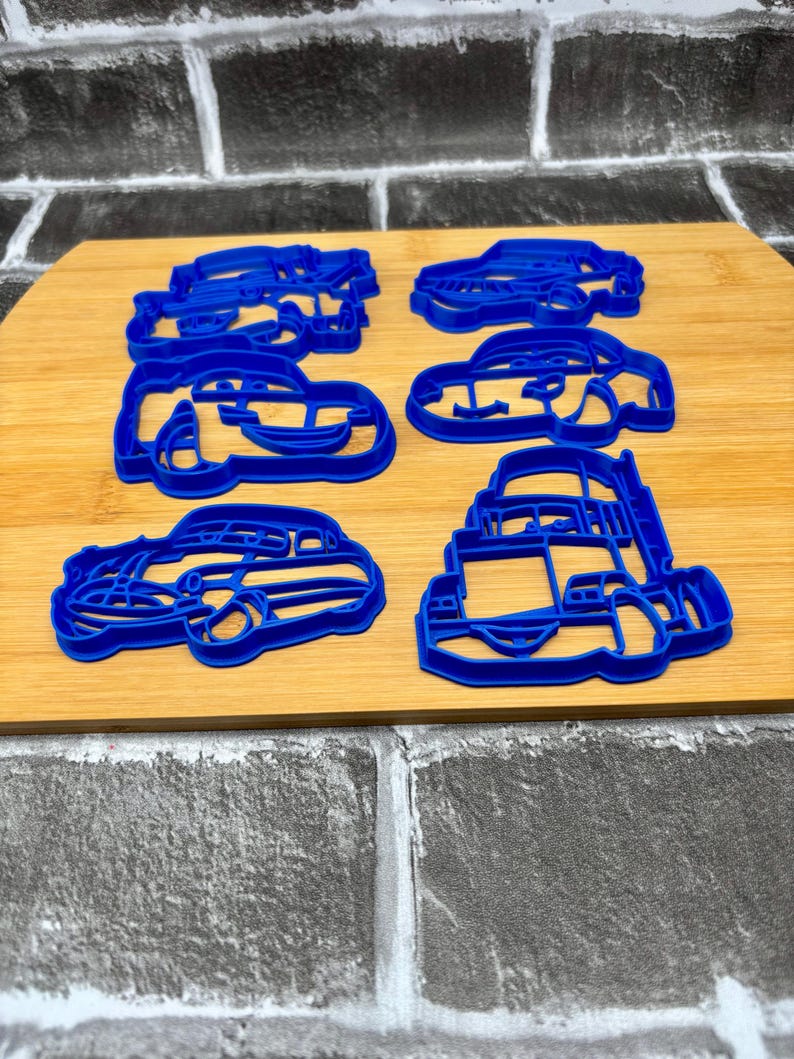 May include: Six blue plastic car-shaped cookie cutters on a wooden surface. The cutters are in various car designs, including a sedan, a convertible, and a truck. The cutters are made of a durable plastic material.