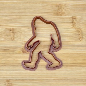 Pode incluir: Um cortador de biscoitos de plástico castanho em forma de Bigfoot, ou Sasquatch, a andar. O cortador tem um contorno detalhado do corpo da criatura, incluindo os braços, pernas e pés. O cortador está colocado numa superfície de madeira.