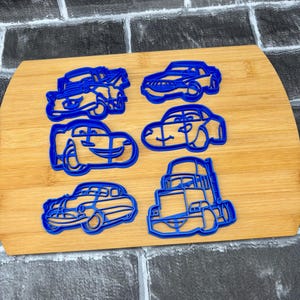 May include: Six blue plastic cookie cutters in various car shapes are arranged on a wooden cutting board. The cutters include a classic car, a semi-truck, and several modern car designs. The background is a gray and black brick pattern.