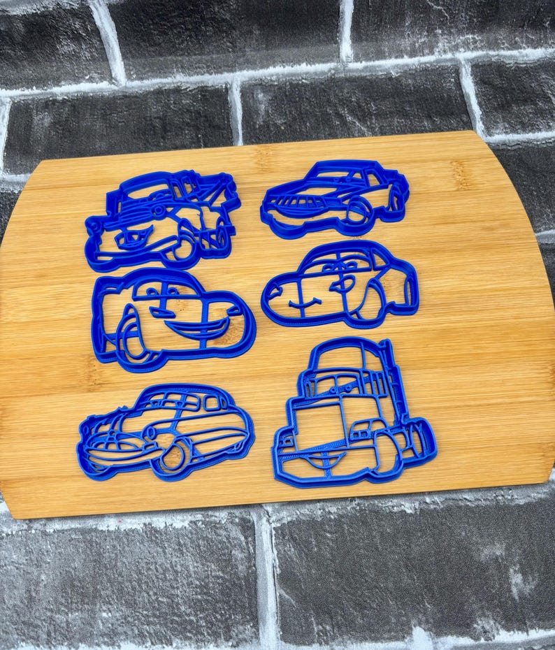 May include: Six blue plastic cookie cutters in various car shapes are arranged on a wooden cutting board. The cutters include a classic car, a semi-truck, and several modern car designs. The background is a gray and black brick pattern.