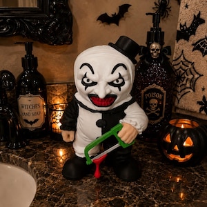 Peut inclure: Une figurine de clown au visage blanc, aux yeux maquill&eacute;s de noir et &agrave; la bouche rouge, portant une tenue noire et blanche et un petit haut-de-forme. La figurine tient une scie &agrave; m&eacute;taux verte avec une goutte rouge suspendue &agrave; la lame.