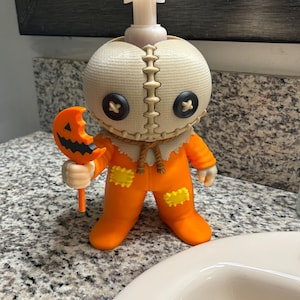 Peut inclure: Un distributeur de savon en forme de personnage avec une t&ecirc;te &agrave; texture de toile de jute, des yeux en boutons noirs et une bouche cousue. La figurine porte une combinaison orange avec des patchs jaunes et tient une sucette citrouille orange.