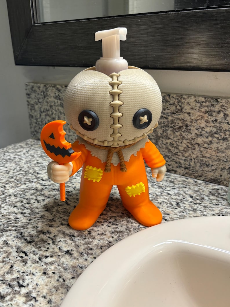 Peut inclure: Un distributeur de savon en forme de personnage avec une t&ecirc;te &agrave; texture de toile de jute, des yeux en boutons noirs et une bouche cousue. La figurine porte une combinaison orange avec des patchs jaunes et tient une sucette citrouille orange.