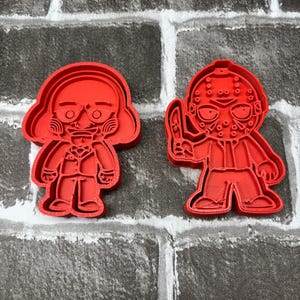 May include: Two red plastic cookie cutters. One is a cartoon figure with a mask and a knife. The other is a cartoon figure with a face and a bow tie. The background is a gray brick wall.