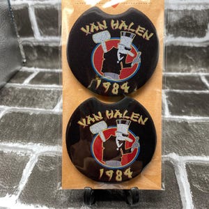 May include: Two black car coasters featuring the Van Halen logo and the year 1984. The coasters have a circular shape with a curved indentation. The logo includes a cartoon figure with a hammer and top hat.