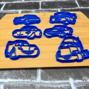 May include: Six blue plastic car-shaped cookie cutters arranged on a wooden cutting board. The cutters feature various car designs, including a race car and a bus. The cutters are made of a bright blue plastic.