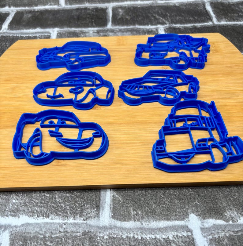 May include: Six blue plastic car-shaped cookie cutters arranged on a wooden cutting board. The cutters feature various car designs, including a race car and a bus. The cutters are made of a bright blue plastic.