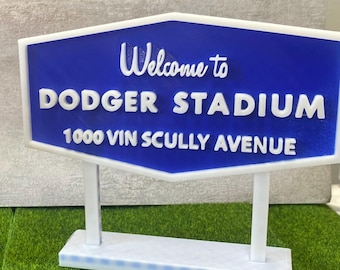 May include: A white and blue sign that reads "Welcome to Dodger Stadium 1000 Vin Scully Avenue". The sign is in a hexagonal shape and is supported by two white posts and a white base. The background is green.