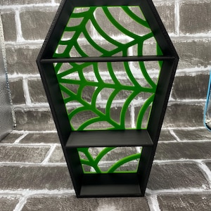 May include: Black coffin-shaped shelf with two shelves and a green spiderweb design. The shelf is made of a solid material and is designed to be mounted on a wall. Ideal for Halloween or gothic decor, it measures approximately 30 cm in height.