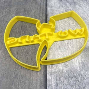Puede incluir: Un cortador de galletas de plástico amarillo con la forma del logotipo de Wu-Tang Clan. La palabra "Wu-Tang" está escrita en el centro del logotipo. El cortador está hecho de una sola pieza de plástico y está diseñado para cortar masa u otros materiales blandos.