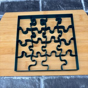 Puede incluir: Un cortador de galletas con forma de rompecabezas de color verde oscuro hecho de plástico. El cortador se coloca sobre una tabla de cortar de madera de color marrón claro. El rompecabezas está formado por 9 piezas individuales.
