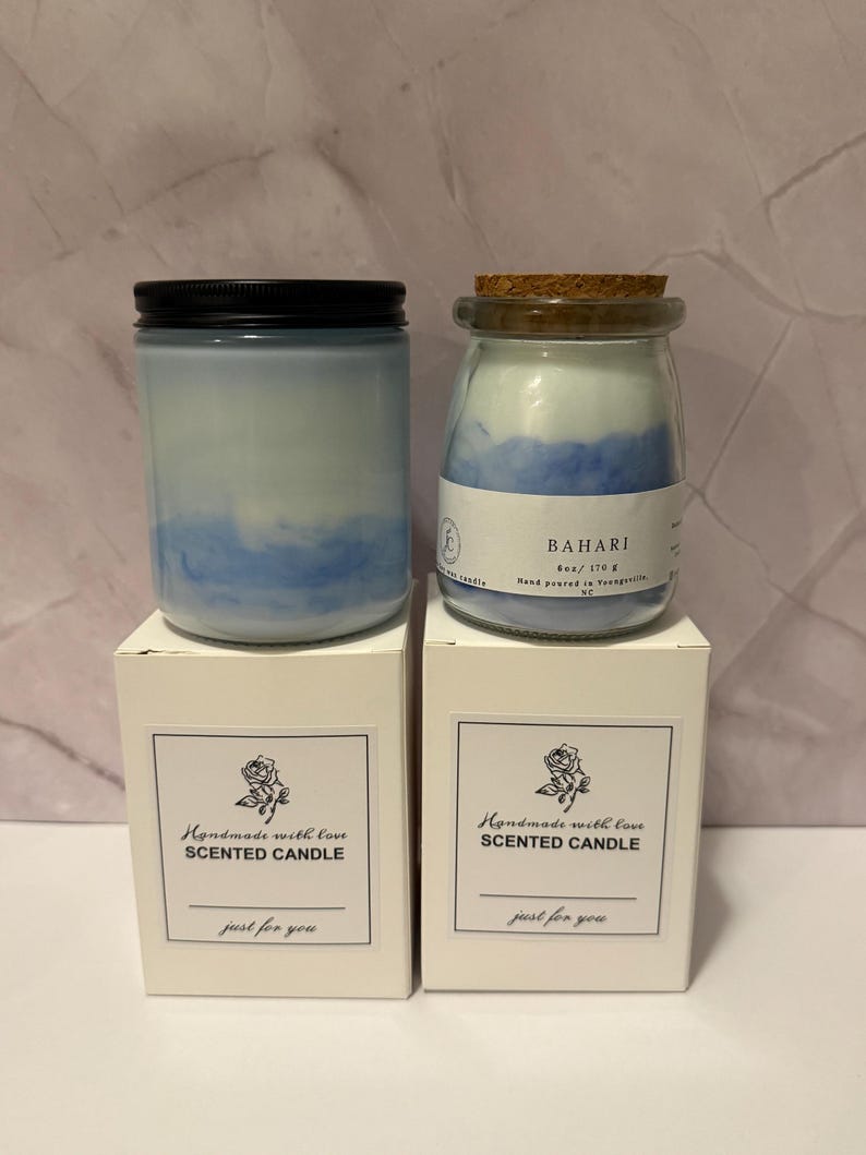 K&ouml;nnte beinhalten: Zwei Duftkerzen in Gl&auml;sern mit blauem und wei&szlig;em Wachs, eine mit schwarzem Deckel und die andere mit Korken. Die Gl&auml;ser stehen auf wei&szlig;en Schachteln mit dem Text "Handmade with love Scented Candle just for you."