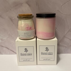 K&ouml;nnte beinhalten: Zwei Duftkerzen in Gl&auml;sern mit rosa und wei&szlig;em Wachs, eine mit Korkdeckel und die andere mit schwarzem Deckel. Die Kerzen stehen auf wei&szlig;en Schachteln mit der Aufschrift "Handmade with love SCENTED CANDLE" und "just for you."