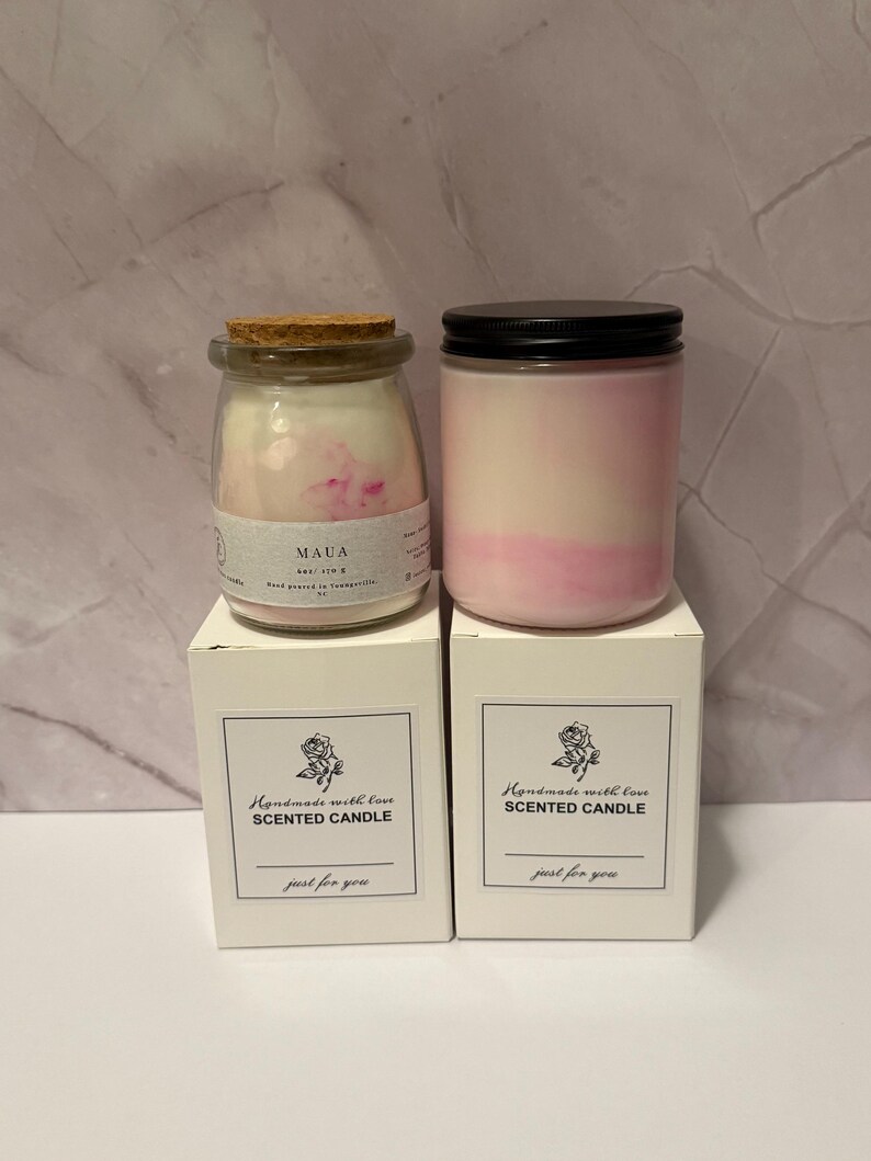 K&ouml;nnte beinhalten: Zwei Duftkerzen in Gl&auml;sern mit rosa und wei&szlig;em Wachs, eine mit Korkdeckel und die andere mit schwarzem Deckel. Die Kerzen stehen auf wei&szlig;en Schachteln mit der Aufschrift "Handmade with love SCENTED CANDLE" und "just for you."