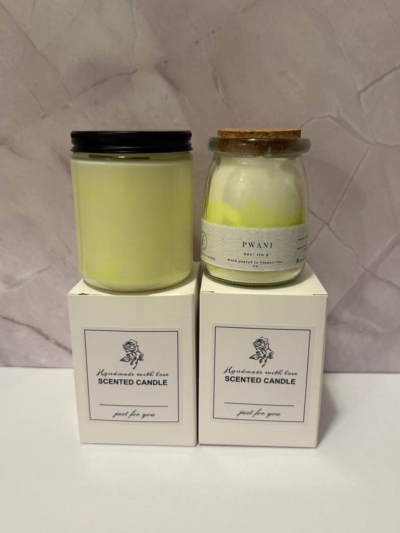 K&ouml;nnte beinhalten: Zwei Duftkerzen in Gl&auml;sern mit schwarzen Deckeln und Korken, auf wei&szlig;en Schachteln. Auf den Schachteln steht "Handmade with love SCENTED CANDLE" und "just for you". Die Kerzen sind gelb.