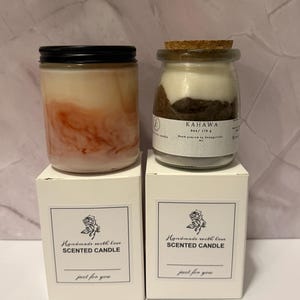 K&ouml;nnte beinhalten: Zwei Duftkerzen in Gl&auml;sern mit schwarzen Deckeln und Korken, auf wei&szlig;en Schachteln. Eine Kerze hat ein geschichtetes rosa und wei&szlig;es Aussehen, die andere ein geschichtetes braunes und wei&szlig;es Aussehen. Auf den Schachteln steht "Handmade with love SCENTED CANDLE".
