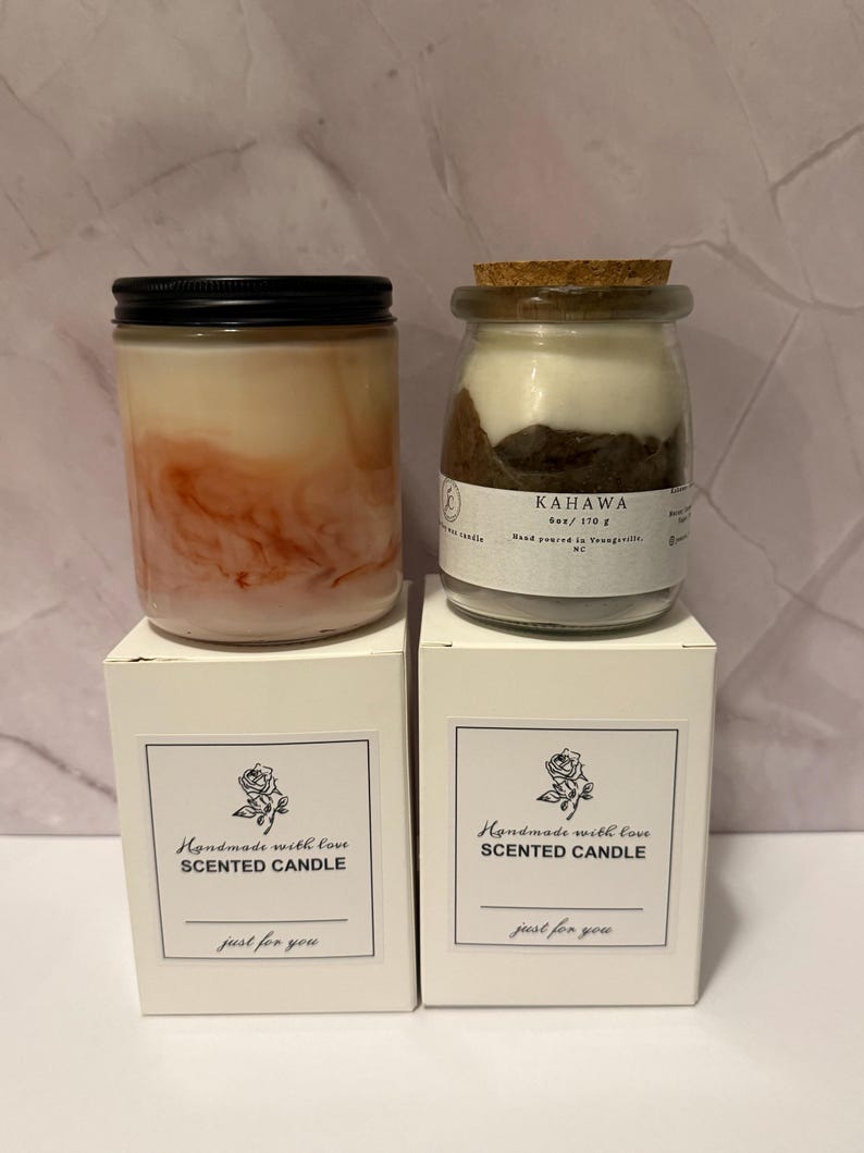 K&ouml;nnte beinhalten: Zwei Duftkerzen in Gl&auml;sern mit schwarzen Deckeln und Korken, auf wei&szlig;en Schachteln. Eine Kerze hat ein geschichtetes rosa und wei&szlig;es Aussehen, die andere ein geschichtetes braunes und wei&szlig;es Aussehen. Auf den Schachteln steht "Handmade with love SCENTED CANDLE".