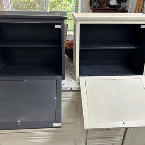 May include: Two rectangular wooden wall cabinets with open shelving and drop-down doors. One cabinet is painted matte black, and the other is painted off-white. Each unit features a single interior shelf and a hinged front panel with metal clips.