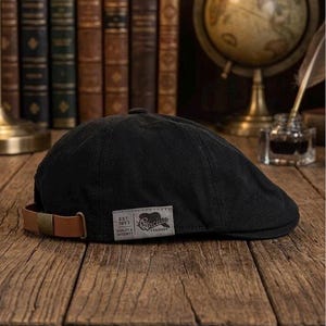 May include: A black flat cap with a brown leather strap and a label. The label says "Est. 1973 Quality & Integrity Cincin Fellows". The cap sits on a wooden surface, with books and a globe in the background.