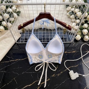 May include: A crochet halter top featuring a cream-colored knit with light blue trim and small yellow flower accents. The garment includes white padded cups, a tie-back closure with long white strings, and a halter neck strap for support.