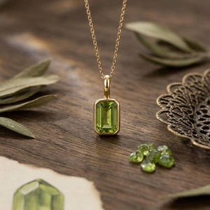 May include: A gold necklace with a rectangular, light green peridot pendant. The pendant is set in a gold bezel and hangs from a delicate gold chain. Several loose peridot stones are scattered nearby on a wooden surface.