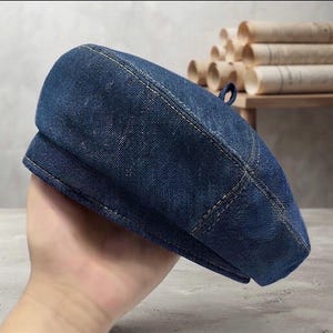 Puede incluir: Una boina de mezclilla azul oscuro con una corona estructurada y un pequeño lazo de tela en la parte superior central. El gorro está confeccionado con tela de mezclilla de algodón resistente con costuras claras visibles y una banda reforzada.