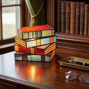 May include: A stack of four stained glass book-shaped boxes in shades of red, orange, yellow, green, and blue. The boxes are on a dark wooden desk, with a window and a bookshelf in the background.
