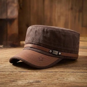 May include: A dark brown military-style cadet cap featuring a flat top and a structured crown. The cap includes a smooth brown leather-like band across the front with a metallic buckle detail and a matching brown brim with a stitched star emblem.