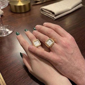 May include: Two gold-toned square ring watches featuring white dials with black Roman numerals and black hands. Each ring has a metallic gold finish and a square-shaped watch face integrated into the band, worn on the fingers of two different hands.