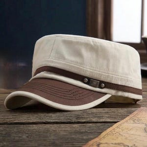 May include: A military-style cadet cap featuring a cream-colored crown and a dark brown brim. A dark brown horizontal band wraps around the base of the crown, accented with a small metallic rectangular detail on the side of the cap.