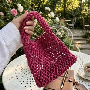 Könnte beinhalten: Eine magenta-farbene Tragetasche mit offenem Gewebe, rechteckiger Form und einem stabilen Griff. Die Tasche besteht aus einem dicken, gewebten Material. Die Tasche wird vor einem verschwommenen Hintergrund eines Gartens gehalten.