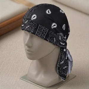 Op de afbeelding: Een zwart-witte bandana-hoofddoek met een klassiek paisley-patroon. De hoofdbedekking heeft een brede band over het voorhoofd en wordt aan de achterkant gestrikt, met een herhalend wit druppel- en krulontwerp op een effen zwarte stof.