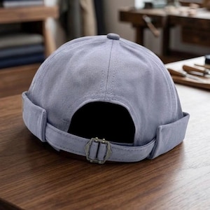 May include: A light blue brimless cap featuring a rounded crown with stitched panels and a rolled edge. The back includes an adjustable strap with a metallic buckle closure, providing a secure fit for this structured headwear accessory.