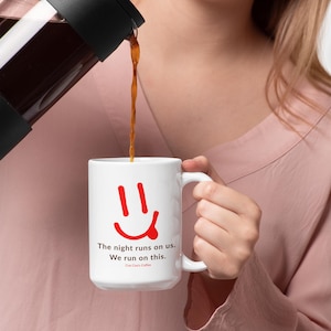 May include: A white ceramic mug with a red smiley face and the text "The night runs on us. We run on this." Coffee is being poured from a black French press. The mug has a white handle.